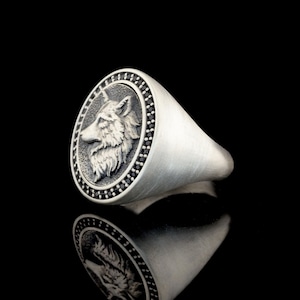 May include: A silver ring with a wolf head design. The wolf head is surrounded by a circle of black stones.