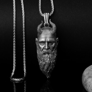 May include: A silver-colored pendant necklace featuring a detailed, sculpted head with horns and a long beard. The pendant is suspended from a matching chain, set against a dark background. The pendant's design is highly detailed.