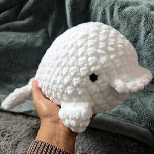 May include: A handmade, white crochet whale plushie with a textured surface. The whale has a rounded body, small fins, and a tail. It features a black eye and is held in a person's hand.