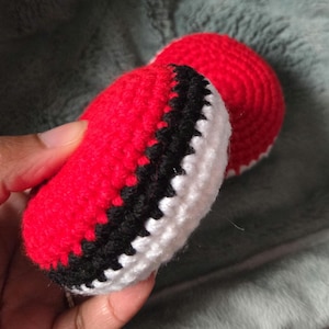 May include: Crocheted red and white object with a black stripe. The object is round and appears to be handmade. The item is held in a hand against a soft, gray background. The crochet work is visible.