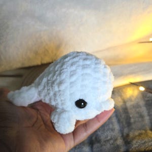 May include: A small, white, crocheted beluga whale plush toy made from soft, textured yarn. The creature features a rounded head, a small tail, and a single visible black plastic eye on its side, giving it a simple and distinct appearance.