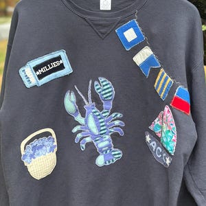 Puede incluir: Sudadera gris oscuro con parches de temática náutica, incluyendo una langosta azul y motivos de banderas. Un parche dice "MILLIES". La sudadera está colgada en una percha de madera.