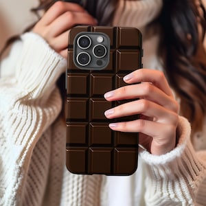 Può includere: Una custodia per telefono marrone scuro con un design a forma di barretta di cioccolato. La custodia ha una superficie strutturata con un motivo a griglia, che ricorda l'aspetto di una barretta di cioccolato. La custodia del telefono ha un ritaglio per la fotocamera con tre obiettivi.