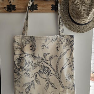 Puede incluir: Un bolso tote beige con un estampado botánico azul oscuro de granadas y follaje. El bolso tiene dos asas y una pequeña etiqueta. Un sombrero de paja cuelga cerca en un perchero de madera con ganchos de metal.