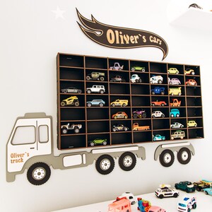 May include: A wall-mounted wooden display shelf shaped like a truck, featuring a rectangular grid with many compartments holding small toy cars. Above the shelf, a wooden sign reads "Oliver's cars," and the truck cab is labeled "Oliver's track."