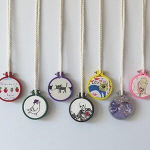 May include: Seven colourful embroidery hoop ornaments with various designs, including a cat, a bird, a strawberry, a skeleton, a fish, and a floral pattern. The hoops are hanging from white cords.