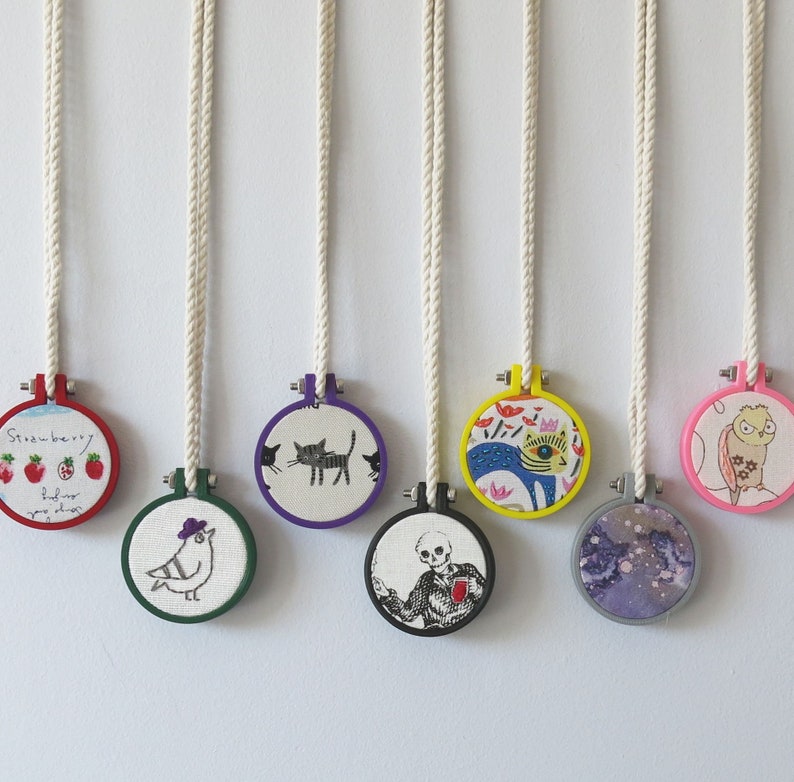 May include: Seven colourful embroidery hoop ornaments with various designs, including a cat, a bird, a strawberry, a skeleton, a fish, and a floral pattern. The hoops are hanging from white cords.