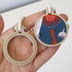 May include: Two embroidery hoops, one with a beige frame and a white fabric insert, and the other with a beige frame and a blue fabric insert with a red bird and floral design.