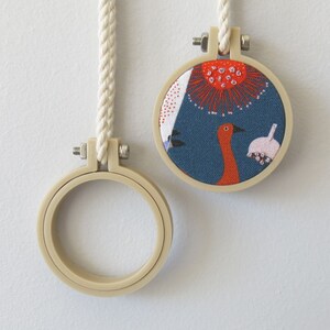 May include: Two embroidery hoops, one with a white cord and a blue fabric with a red and white floral design and a bird, and the other with a white cord and a blank hoop.