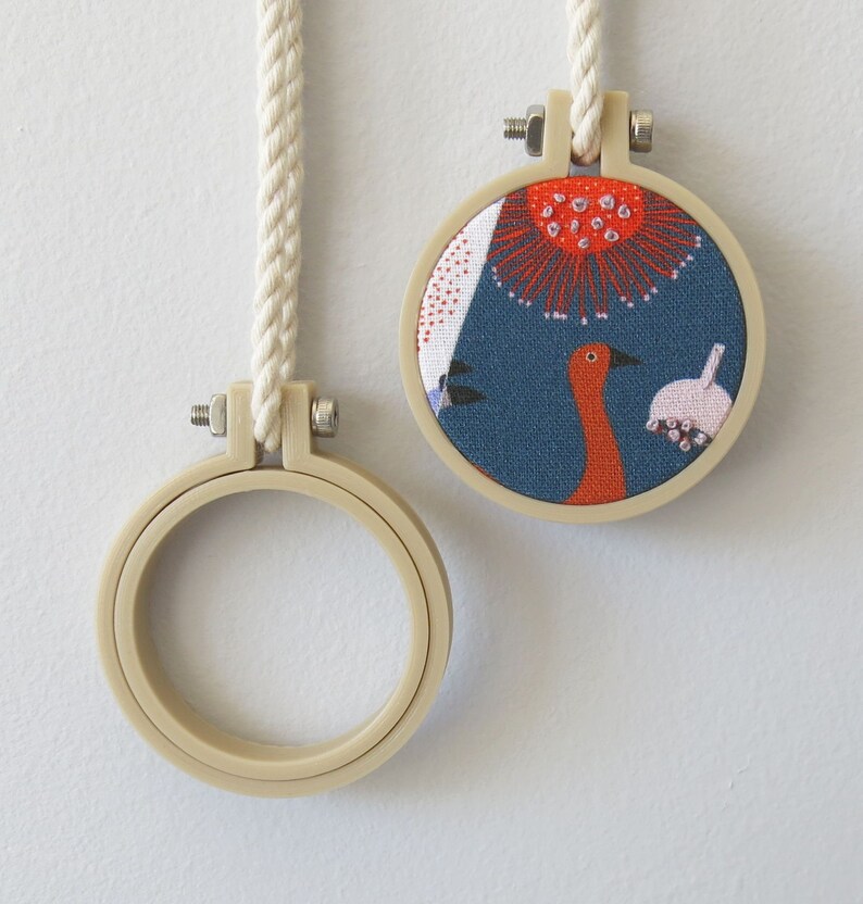 May include: Two embroidery hoops, one with a white cord and a blue fabric with a red and white floral design and a bird, and the other with a white cord and a blank hoop.