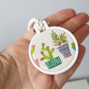 May include: A white embroidery hoop with a white background and colourful embroidery of potted plants. The plants include a cactus with pink flowers and a leafy plant with pink flowers.