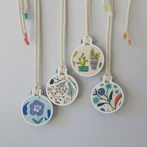 May include: Five white embroidery hoops with colourful floral designs. Each hoop is attached to a white cord and hangs from a white surface.