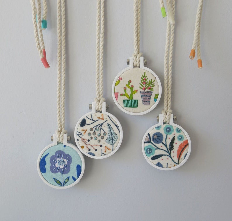 May include: Five white embroidery hoops with colourful floral designs. Each hoop is attached to a white cord and hangs from a white surface.