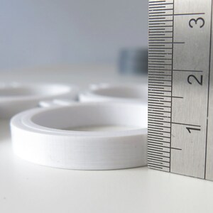 May include: A white plastic ring measuring 2.5 cm in diameter, next to a metal ruler marked with centimetres.
