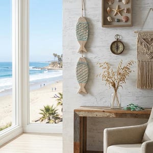 May include: A vertical wall hanging featuring two carved wooden fish shapes wrapped in light blue and white netting, suspended by a rope. The fish are displayed on a white textured wall alongside a framed collection of seashells and a round compass.