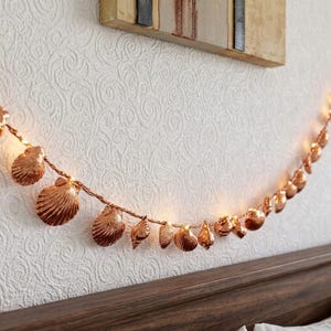 May include: A string of decorative lights featuring metallic copper-coloured seashell covers. The shells are arranged along a thin wire, with small warm-toned lights glowing from within each individual shell, creating a soft illumination effect.