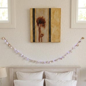May include: A string of decorative lights featuring small, pale, shell-like accents strung along a thin wire. The lights emit a soft, cool-toned glow, creating a gentle arc across the wall below a rectangular painting of a brown palm tree.