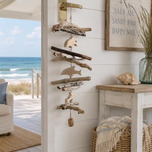 May include: A driftwood mobile featuring wooden fish and geometric shapes hangs on a white wall. The mobile is suspended by a gold-coloured handle. A wooden sign with the text "SALT AIR SANDY HAIR HAPPY SOUL" is visible.