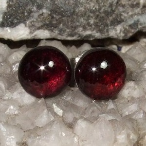 May include: A pair of round, deep red garnet stud earrings. The polished gemstones have a high lustre and are set against a backdrop of white quartz crystals and grey rock.