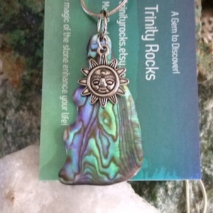 Puede incluir: Un collar de plata con un dije de sol y un dije de concha de abulón verde, azul y morado. El dije tiene forma de lágrima y tiene un brillo iridiscente natural. El collar se muestra sobre una superficie blanca con una tarjeta de presentación que dice "A Gem to Discover! Trinity Rocks Trinityrocks.etsy.com The magic of the stone enhance your life!"