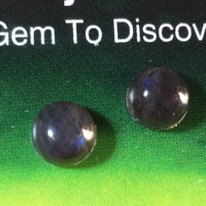 May include: Two round, dark blue labradorite gemstones with iridescent flashes of colour. The gemstones are on a card that reads "Trinity Rocks A Gem To Discover!" and "Labradorite Newfoundland".