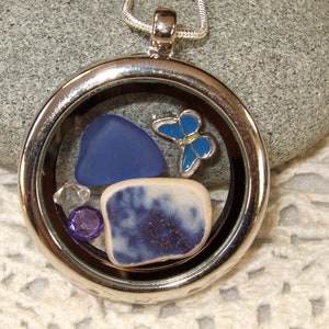 May include: A silver locket necklace with a glass insert containing a blue butterfly charm, a blue sea glass piece, a white and blue ceramic tile, and two clear and purple rhinestones.