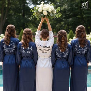 Può includere: Una sposa e le sue damigelle in abiti blu navy e bianchi, con la sposa che tiene in mano un bouquet di fiori bianchi. L'abito della sposa è bianco con "Victoria Bride" stampato sul retro. Gli abiti delle damigelle sono blu navy.
