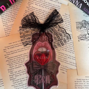 May include: A decorative sticker featuring a red heart-shaped lollipop with the word "VILLAIN" on it, framed by a dark bow and the word "ERA". The sticker is adorned with a black lace bow and set against a backdrop of vintage book pages.