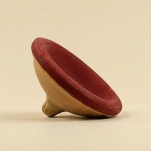 Op de afbeelding: Een houten tol met een rode rand en een natuurlijke houten body. Het speelgoed staat iets schuin, wat de conische vorm en de afgeronde basis laat zien. De achtergrond is effen en neutraal.