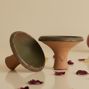 Op de afbeelding: Twee terracotta-kleurige keramische objecten met groene accenten, één rechtop en één op zijn kant. Een rode granaatappel en verspreide bloemblaadjes zijn ook zichtbaar. De objecten hebben een paddenstoelachtige vorm.