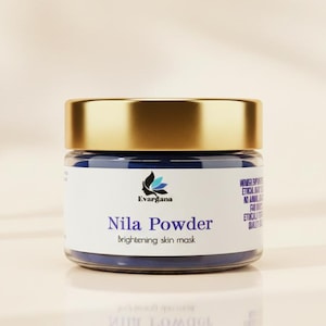 Op de afbeelding: Een klein glazen potje met een gouden deksel en een blauw etiket. Op het etiket staat "Nila Powder brightening skin mask" met een logo. Het potje is gevuld met blauw poeder. De achtergrond is een zachte, neutrale kleur.
