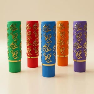 Könnte beinhalten: Fünf farbige Lippenstifttuben. Jede Tube hat eine andere Farbe: grün, rot, blau, orange und lila. Die Tuben haben ein goldenes Schmetterlingsmuster und goldene Akzente. Die Lippenstifttuben stehen aufrecht.