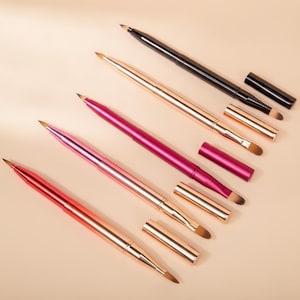 May include: Four retractable make-up brushes with metallic barrels in black, rose gold, magenta, and red. Each brush features a pointed synthetic tip and a matching removable cap, arranged diagonally on a solid light beige surface for display.