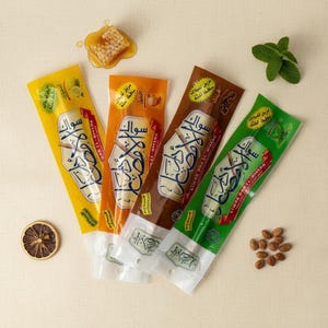 May include: Four brightly coloured Siwak stick packages, each with Arabic text and flavour indicators. The packages are yellow, orange, brown, and green. A piece of honeycomb, a sprig of mint, a dried lemon slice, and a small pile of nuts are also present.