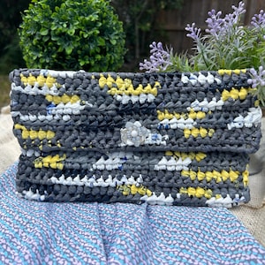 May include: A handmade clutch with a crocheted design in shades of gray, yellow, and white. The clutch features a decorative clasp and is placed on a patterned fabric. The bag is a stylish accessory.