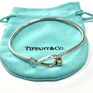 May include: A polished sterling silver bangle bracelet featuring a fish-shaped charm with a small spherical bead at the end. The bracelet is resting on a light blue drawstring pouch with the text Tiffany & Co. printed in black on the front surface.