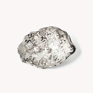 Peut inclure: Une broche argentée de forme organique, irrégulière et texturée. La surface présente une finition rugueuse et cratérisée avec quatre accents sphériques lisses et polis répartis sur la face de la pièce, créant un contraste de texture.