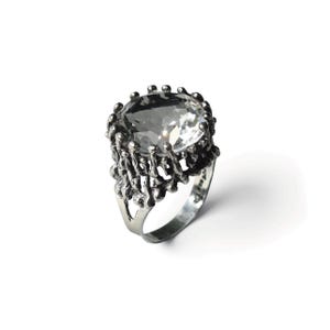 Könnte beinhalten: Ein silberfarbener Ring mit einem großen, klaren, facettierten ovalen Edelstein, der in einem strukturierten, organisch wirkenden Metallkäfig gefasst ist. Die Ringschiene ist glatt und poliert und bildet einen Kontrast zur komplexen Metallarbeit.