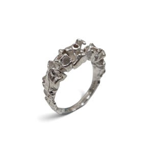 May include: A silver ring featuring a textured, organic band with an uneven, molten-like surface. The metal has a matte, metallic finish with irregular protrusions and indentations along the top edge, creating a rugged and sculptural appearance.