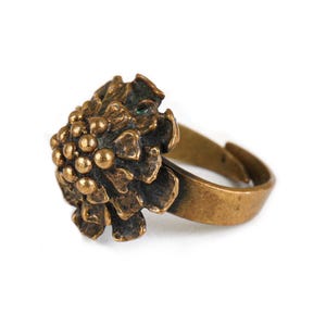 Könnte beinhalten: Ein bronzefarbener Metallring mit einem dreidimensionalen Blumendesign. Das Mittelstück ist wie eine Blume geformt, mit einer texturierten, abgerundeten Ansammlung kleiner Kugeln in der Mitte, umgeben von geschichteten, unregelmäßigen Blütenblättern. Der Ring ist verstellbar.