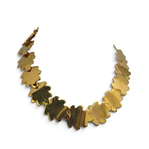 Op de afbeelding: Een goudkleurige metalen ketting bestaande uit een reeks verbonden, platte, bladvormige schakels die in een gebogen, halfronde formatie zijn gerangschikt. Elke individuele schakel heeft een glad, reflecterend oppervlak en een geschulpte rand.