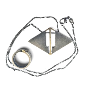 Könnte beinhalten: Eine dunkelgraue Metallkette mit einem geometrischen rautenförmigen Anhänger mit einem vertikalen messingfarbenen Stab, begleitet von einem passenden dunkelgrauen Metallring mit einem messingfarbenen Akzent. Die Kette enthält eine dünne Kette.