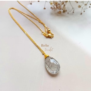 May include: A gold-colored chain necklace with a teardrop-shaped pendant. The pendant is a clear stone with black inclusions. The necklace is displayed on a white surface. The text "Belo Trinket" is visible.