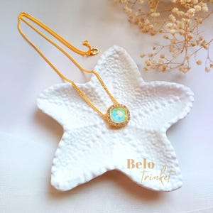 Op de afbeelding: Een goudkleurige ketting met een vierkante, iriserende hanger omringd door kleine, heldere kristallen. De ketting ligt op een witte, stervormige schaal. De tekst "Belo Trinket" is zichtbaar.