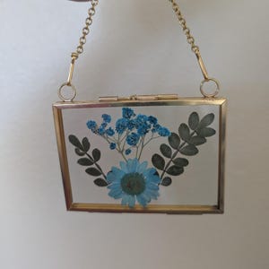 May include: A rectangular, gold-framed glass display case with a gold chain handle. The case contains a blue flower arrangement with green leaves. The arrangement includes a large blue daisy-like flower and smaller blue flowers.