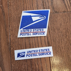 May include: Two embroidered patches featuring the United States Postal Service logo. The top patch is a trapezoid with a blue eagle graphic and the text UNITED STATES POSTAL SERVICE. The bottom patch is a rectangle with the same logo and text.