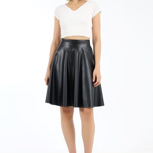 May include: A woman wearing a white, short-sleeved, V-neck crop top and a black, faux leather, flared midi skirt. She is also wearing black loafers with silver hardware. The outfit is set against a white background.