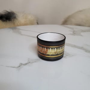 May include: A small, cylindrical ceramic container. It has a black exterior and a white interior. The exterior features horizontal bands of brown, yellow, and white, with a decorative pattern. The container sits on a white marble-patterned surface.