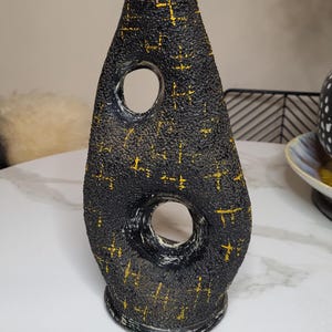 May include: Black and gold ceramic vase with a textured surface and two circular cutouts. The vase has a unique, abstract shape.