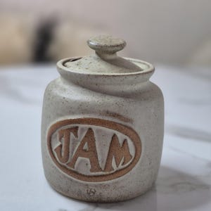 May include: A beige ceramic jam jar with a lid. The jar has a brown oval with the word "JAM" in brown letters. The jar has a small knob on the lid and a speckled finish.